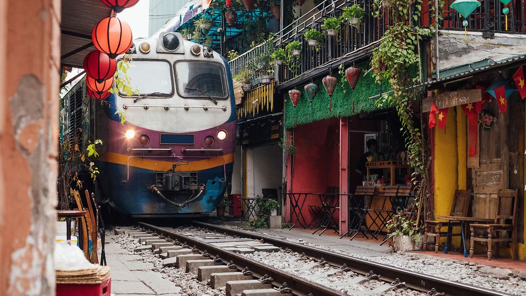 Train travelling through Hanoi, in the famous tourist location. It shows the extreme proximity to the shops and buildings on either side. Traditional colourful lanterns and flags decorate the buildings and cafes en route.